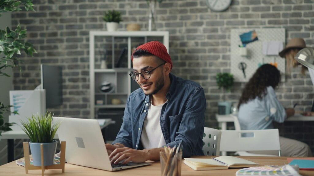 A young man in a beanie working diligently on a laptop in a stylish, modern office space.