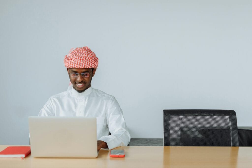 Middle Eastern man in traditional attire using a laptop at his office desk, smiling while working.