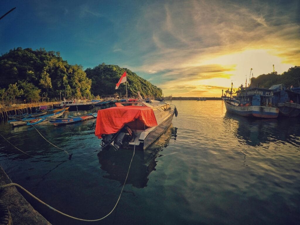 A stunning sunset view at Girisubo Harbor, Indonesia, with boats and lush hills.