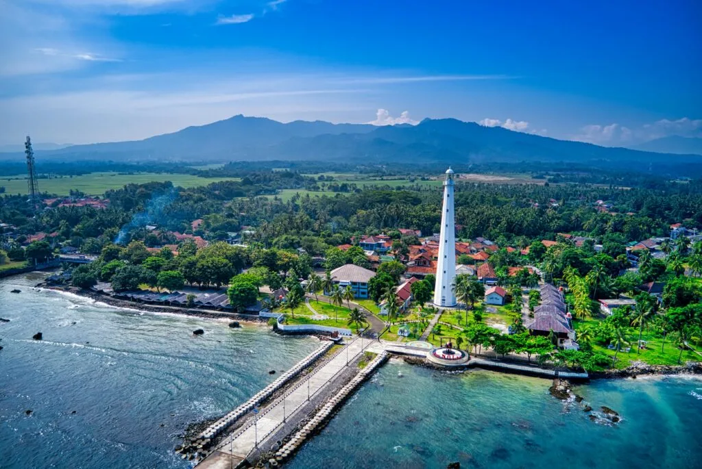 Stunning aerial view of the lighthouse and coastline in Banten, Indonesia, surrounded by lush greenery and mountains.