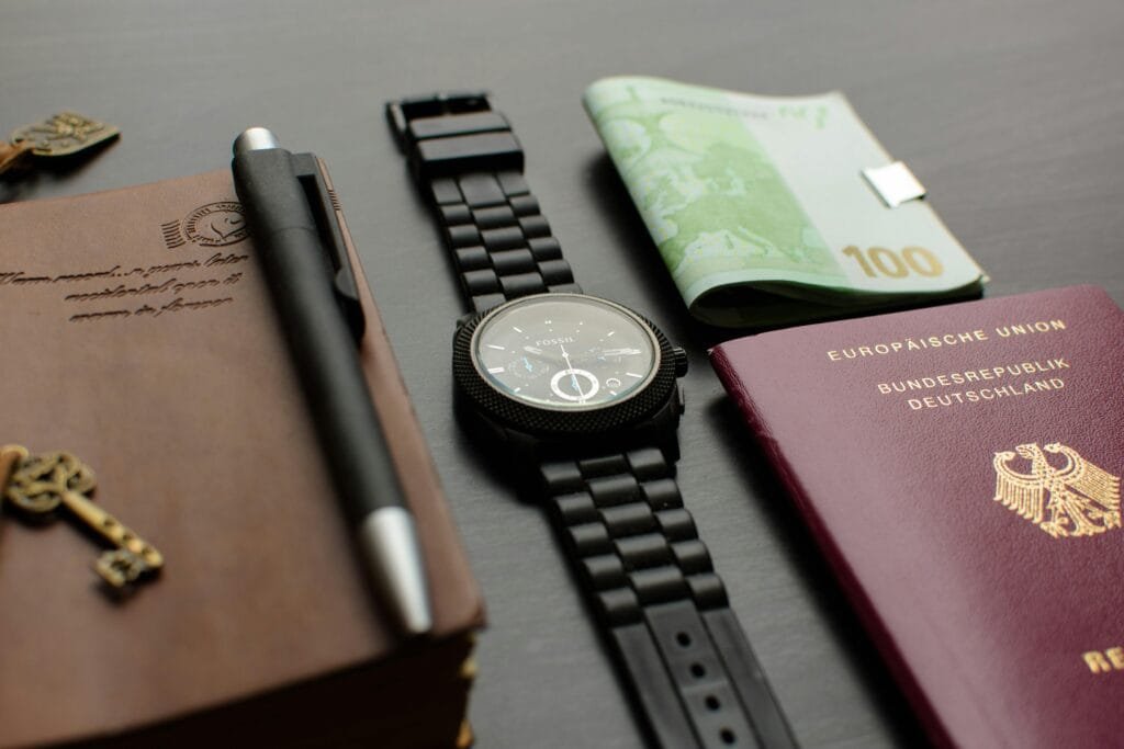 A neatly arranged flat lay depicting essential travel items such as a German passport, wristwatch, and euro bills on a desk.